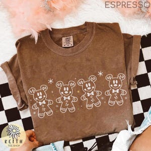 May include: A brown short-sleeve t-shirt featuring a white graphic print of four gingerbread-style Mickey Mouse characters arranged in a horizontal row, accented with small white snowflakes and stars, with the word ESPRESSO printed at the top.