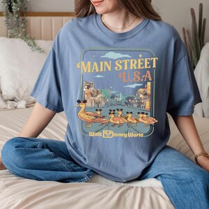 May include: A blue t-shirt with a Main Street U.S.A. design, featuring a duck family wearing Mickey Mouse ears. The shirt has a retro style with the words "Walt Disney World" at the bottom.