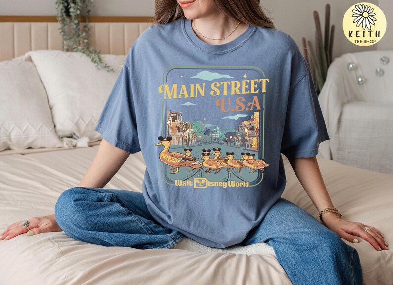 May include: A blue t-shirt with a Main Street U.S.A. design, featuring a duck family wearing Mickey Mouse ears. The shirt has a retro style with the words "Walt Disney World" at the bottom.