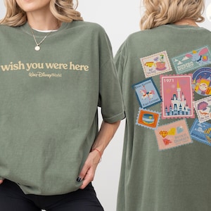 May include: Olive green t-shirt with the text "wish you were here" and "Walt Disney World" printed on the front. The back features a collection of colorful postage stamp designs with Disney-themed illustrations, including a castle and Dumbo.