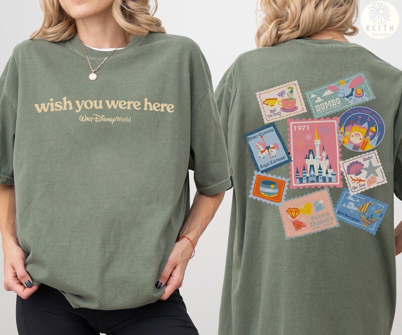 May include: Olive green t-shirt with the text "wish you were here" and "Walt Disney World" printed on the front. The back features a collection of colorful postage stamp designs with Disney-themed illustrations, including a castle and Dumbo.