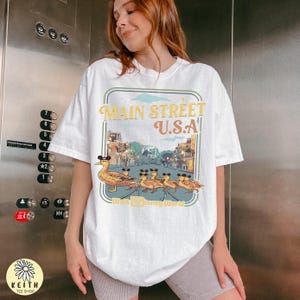 May include: White t-shirt with a Main Street U.S.A. graphic in a vintage style. The design shows a duck in a Mickey Mouse hat leading ducklings, also wearing hats. The words "Walt Disney World" are included in the design.