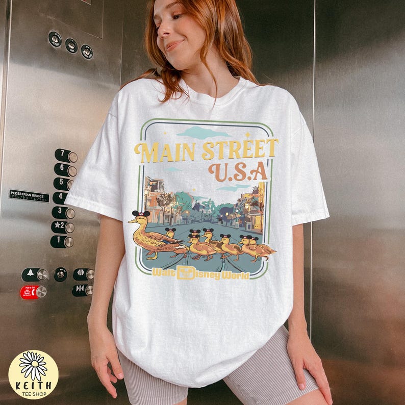 May include: White t-shirt with a Main Street U.S.A. graphic in a vintage style. The design shows a duck in a Mickey Mouse hat leading ducklings, also wearing hats. The words "Walt Disney World" are included in the design.