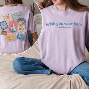 May include: A light purple t-shirt with the text "wish you were here" and "Walt Disney World" printed in blue. The back of the shirt features a collection of colorful postage stamp designs with various illustrations and the year 1971.