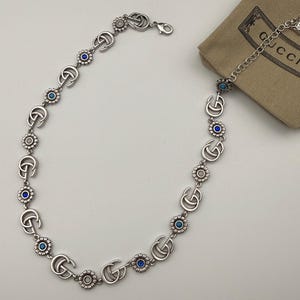 Puede incluir: Collar plateado con logotipos "G" entrelazados y detalles florales con centros azules y blancos. El collar se muestra sobre una superficie blanca, con una pequeña bolsa marrón en el fondo.