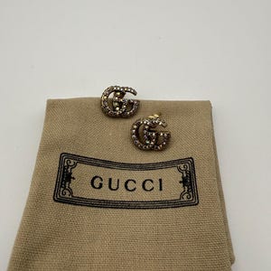 Puede incluir: Pendientes de botón con el logotipo "GG" entrelazado, incrustados con pequeñas piedras brillantes. Los pendientes descansan sobre una bolsa de tela beige con el logotipo "GUCCI" en negro. Fondo blanco.