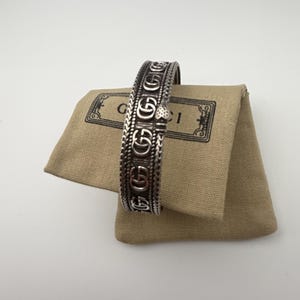 Puede incluir: Brazalete plateado con un patrón repetido del logotipo "G" y detalles intrincados. El brazalete se exhibe sobre una bolsa de tela color beige con el nombre de la marca impreso. La estética general es lujosa y elegante.