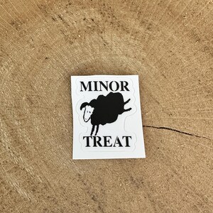 May include: A white sticker with the words "MINOR TREAT" in black, and a black sheep illustration. The sheep is shown with its legs in the air. The sticker is on a wooden surface.