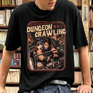 May include: Black t-shirt with a graphic featuring a man driving a vehicle with a cat, and the text "GET IN LOSER WE'RE GOING DUNGEON CRAWLING". The design has a vintage, comic book style.