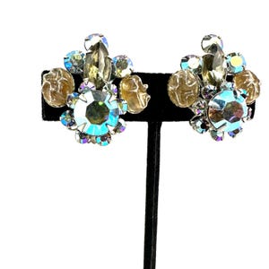 May include: A pair of cluster earrings featuring large iridescent blue-toned central crystals surrounded by smaller clear, light blue, and textured amber-colored stones arranged in a floral-like pattern with a metallic setting.