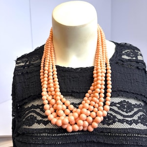 May include: A multi-strand necklace featuring several layers of smooth, round, peach-colored beads. The beads vary slightly in size, creating a graduated effect across the multiple strands that drape around the neck area in a cascading style.