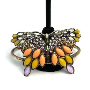 Può includere: Un bracciale rigido in metallo color argento con una grande farfalla centrale. Le ali della farfalla sono decorate con cabochon a forma di marquise nei toni del pesca, giallo e lavanda, accentuati da piccoli strass trasparenti e viola.