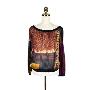May include: A long-sleeve blouse featuring a wide, scalloped neckline and a multi-panel design. The fabric displays a mix of patterns including paisley, geometric shapes, and floral motifs in shades of burgundy, mustard yellow, black, and purple.