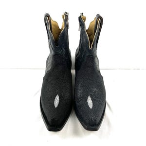 Könnte beinhalten: Ein Paar schwarze Stiefel mit einer texturierten Rochenhaut-Optik am Vorderblatt und einem Schaft aus glattem Leder. Jeder Stiefel hat eine spitz zulaufende Kappe, eine markante weiße ovale Einlage vorne, einen seitlichen Reißverschluss und ein beiges Innenfutter.