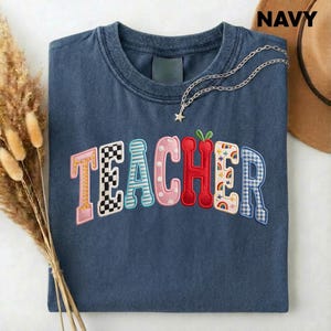 Könnte beinhalten: Marineblaues T-Shirt mit dem Wort "TEACHER" in bunten, gemusterten Buchstaben gestickt. Das Shirt hat einen Rundhalsausschnitt und ist mit einer silbernen Halskette mit Sternanhänger versehen. Ein brauner Hut ist im Hintergrund zu sehen.