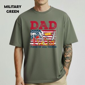 May include: Military green t-shirt with a graphic that says "DAD" in red letters above an American flag design with tools. The text "FIXER OF ALL THINGS" is below the tools.