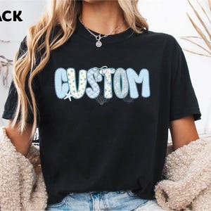 May include: Black t-shirt with the word "CUSTOM" in a light blue, textured font. The letters are decorated with white stars, seashells, and floral patterns. The word "BLACK" is in the top left corner.