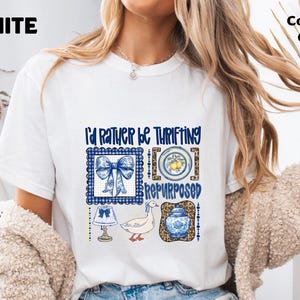 Op de afbeelding: Een wit t-shirt met korte mouwen en een grafische print met de tekst I'd Rather Be Thrifting Repurposed. Het ontwerp bevat illustraties van een blauwe strik, een bord met citroenen, een lamp, een gans en een blauw-witte pot.
