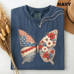 Könnte beinhalten: Marineblaues T-Shirt mit einem Schmetterlingsdesign. Die Schmetterlingsflügel sind mit einem amerikanischen Flaggenmuster und floralen Elementen in Rot, Weiß und Blau verziert. Eine silberne Halskette mit einem Sternanhänger liegt auf dem T-Shirt.