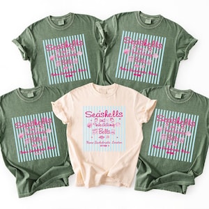 以下が含まれることがあります： オリーブグリーン4枚とピーチ1枚のTシャツ5枚。各シャツには、ピンク色の「Seashells and Wedding Bells」の文字と、青と白のストライプが入った正方形のデザインが施されています。「Name Bachelorette Location」の文字も表示されています。
