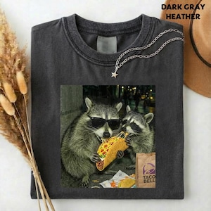 May include: Dark gray heather t-shirt featuring a graphic of two raccoons wearing sunglasses and eating tacos. A silver chain with a star pendant is draped across the shirt. The Taco Bell logo is visible on a brown paper bag.
