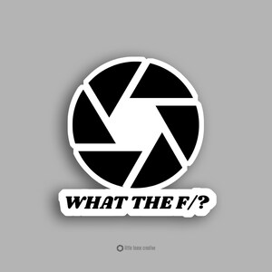 May include: A white sticker featuring a black and white camera aperture design and the text "WHAT THE F/?" on a grey background. The sticker has a white border and a small logo at the bottom.