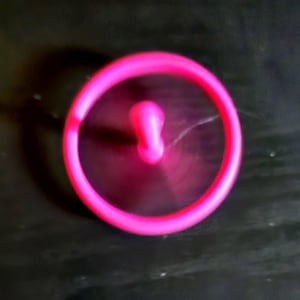 May include: A bright pink spinning toy with a circular outer ring and a central, elongated, rounded piece. The toy is in motion, creating a blurred effect. The background is a dark, solid color.