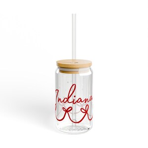 Puede incluir: Un vaso de vidrio transparente con tapa de bambú y pajita transparente. El vaso tiene la palabra "Indiana" en escritura cursiva roja con un diseño de lazo rojo. Diseñado para bebidas frías.