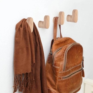 Könnte beinhalten: Hölzerne Wandhaken, an denen ein brauner Schal und ein brauner Lederrucksack hängen. Die Haken sind aus hellem Holz gefertigt und an einer weißen Wand befestigt. Der Schal hat einen Fransenrand.