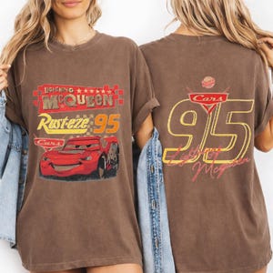 以下が含まれることがあります： 映画「カーズ」のライトニング・マックィーンが描かれた茶色のTシャツ。前面には「Lightning McQueen」、「Rusteze 95」、赤い車のイラスト。背面には黄色と赤で「95」と「Lightning McQueen」がデザインされています。映画をテーマにしたTシャツ。
