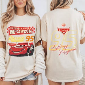 以下が含まれることがあります： 映画「カーズ」のライトニング・マックィーンが描かれたベージュ色のTシャツ。前面には「Lightning McQueen」と「Rusteze 95」の文字とキャラクターのグラフィック。背面には大きな「95」と「Lightning McQueen」の文字とカーズのロゴが描かれています。