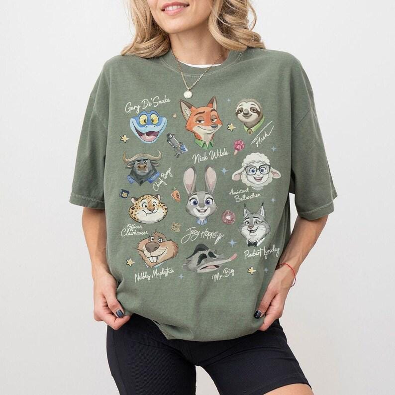 May include: An olive green t-shirt with cartoon character portraits from the film Zootropolis. Characters include Nick Wilde, Judy Hopps, Flash, and others. The shirt has a crew neck and short sleeves.