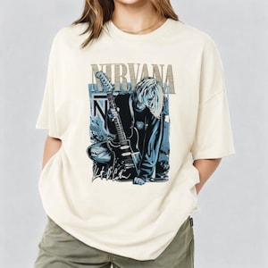 May include: Cream-colored t-shirt featuring a graphic of a person with long hair holding a guitar, with the band name "NIRVANA" above. The design includes blue and black accents and the signature "Kurt Cobain".