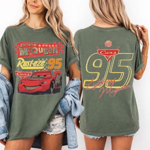 以下が含まれることがあります： 映画「カーズ」のライトニング・マックィーンが描かれたオリーブグリーンのTシャツ。前面には「Lightning McQueen Rusteze 95」の文字と赤い車のイラスト。背面には大きな「95」と「Lightning McQueen」が黄色と赤色で表示されています。