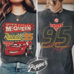 以下が含まれることがあります： 映画「カーズ」のライトニング・マックィーンが描かれた濃いグレーのTシャツ。前面には「Lightning McQueen」、「Rusteze 95」、赤いレーシングカーの文字とイラスト。背面には「Cars 95」と「Lightning McQueen」が表示されています。