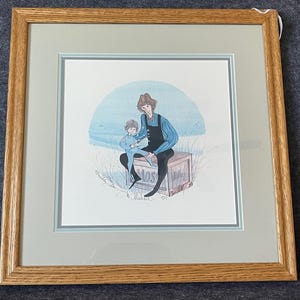 May include: Framed print of a mother and child sitting on a wooden box, with a blue and white background. The artwork features a woman in a blue shirt and black overalls, and a child in a blue outfit. The frame is made of light brown wood.