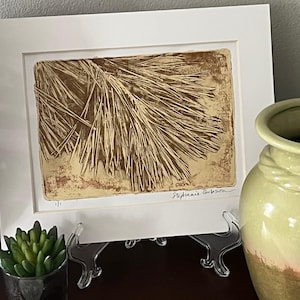 May include: A framed art print featuring a detailed, textured depiction of pine needles in shades of brown and beige. The artwork is signed by Stephanie Gibson and displayed on a clear easel, with a small succulent and a green vase in the background.