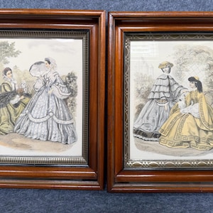 May include: Two rectangular wooden frames containing vintage-style illustrations of figures in historical attire. The left print shows two individuals in outdoor settings, while the right print depicts two figures in layered dresses and hats.