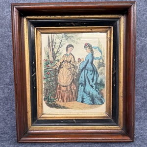 May include: A framed print featuring two figures in Victorian-style attire standing in a garden setting. One figure wears a tan dress, the other a blue dress with a parasol. The frame is constructed of dark wood with gold and black accents.