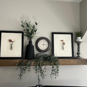 May include: A wooden shelf displays home decor items. Two framed floral prints flank a black vase with greenery and a round clock. A small plant sits on a decorative stand. Trailing faux greenery hangs below the shelf.