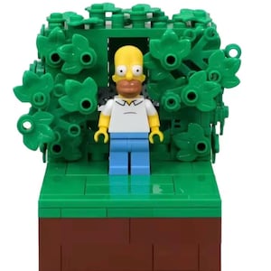 Puede incluir: Una figura de Lego de Homer Simpson se encuentra frente a un telón de fondo verde y frondoso. Homer tiene piel amarilla, una camisa blanca, pantalones azules y un bigote marrón. La base es verde con una sección inferior marrón.