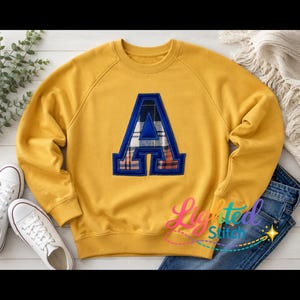 May include: A mustard yellow crewneck sweatshirt featuring a large, central letter A applique. The letter has a blue border and a plaid fabric fill in shades of blue, white, and orange. The text "Lighted Stitch" appears in colorful script.