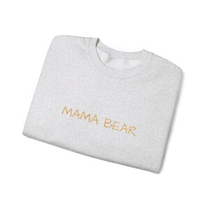 May include: A light gray sweatshirt with the embroidered text "MAMA BEAR" in a gold color. The sweatshirt is folded and has a crew neck.