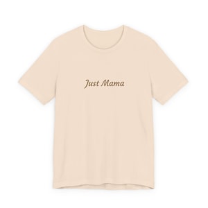 May include: A pale beige t-shirt featuring the words "Just Mama" in a brown script font. The shirt has short sleeves and a crew neck. The t-shirt is made of a soft material.
