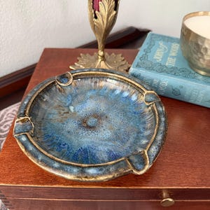 May include: A round ceramic ashtray featuring a variegated glaze in shades of deep blue, light blue, and brown. The ashtray has a wide, textured rim with four distinct notches for resting items, and a glossy finish with a mottled, speckled appearance.