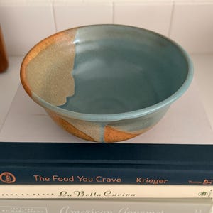 May include: A ceramic bowl with a smooth, light blue interior glaze and a textured, earthy orange and tan exterior finish. The bowl sits atop a stack of three books titled The Food You Crave, La Bella Cucina, and American Gourmet.