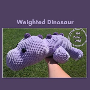 Könnte beinhalten: Ein hellviolettes gehäkeltes Dinosaurier-Spielzeug mit strukturierter Oberfläche, dunklen runden Augen und kleinen violetten Stacheln auf dem Rücken. Der Text "Weighted Dinosaur" und "PDF Pattern Only!" weist darauf hin, dass dies eine digitale Datei ist.