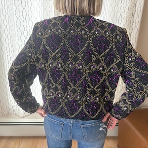 Peut inclure: Veste noire ornée de perles dorées et violettes. Le motif présente un motif détaillé de formes et d'ornements reliés. La veste à manches longues est portée sur un jean bleu.