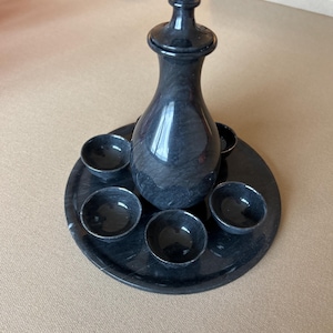 Könnte beinhalten: Ein schwarzes Marmor-Likör-Set mit einer Karaffe mit Deckel, einem runden Tablett und sechs kleinen Schalen. Das Set besteht aus poliertem schwarzen Marmor mit subtilen Maserungen, was eine anspruchsvolle und elegante Ästhetik schafft.