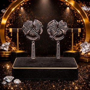 Peut inclure: Une paire de boucles d'oreilles argent&eacute;es en forme de fleur avec des accents de pierres noires. Chaque boucle d'oreille pr&eacute;sente un motif floral d&eacute;taill&eacute; avec un &eacute;l&eacute;ment rectangulaire pendant. Les boucles d'oreilles sont expos&eacute;es sur un pr&eacute;sentoir &agrave; bijoux en velours noir.